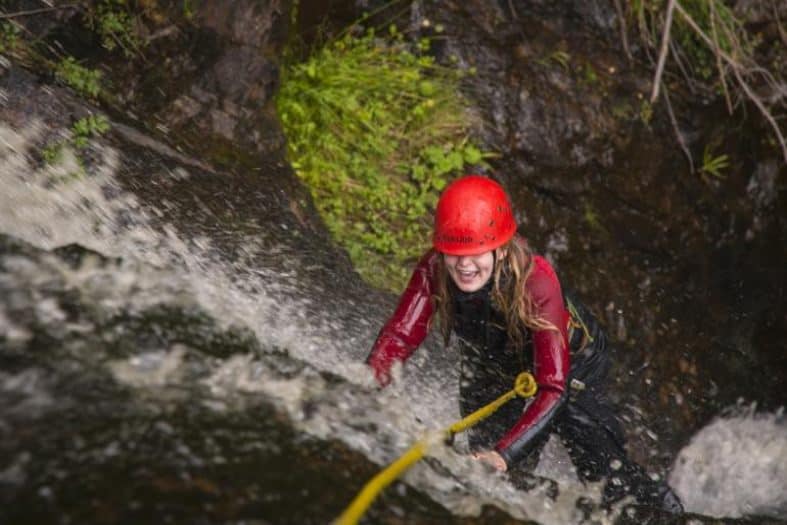 A young girl in a red wetsuit climbs up a fast-flowing waterfall with a big smile on her face. A yellow rope is guiding her and her red hard hat is covering her eyes as she  gets completely drenched from the water on her teens adventure holiday